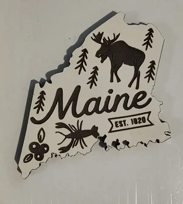 White Maine state–shaped wooden magnet with laser-engraved moose, pine trees, lobster, blueberries, and “Maine Est. 1820” design