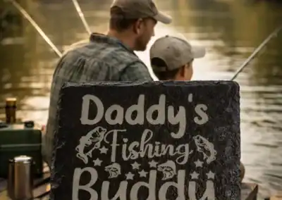 Daddy’s Fishing Buddy slate coaster on wooden table with iced tea and cranberry juice, father and son fishing on a lake in background