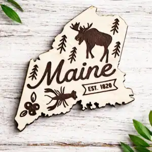 White Maine state–shaped wooden magnet with laser-engraved moose, pine trees, lobster, blueberries, and “Maine Est. 1820” design