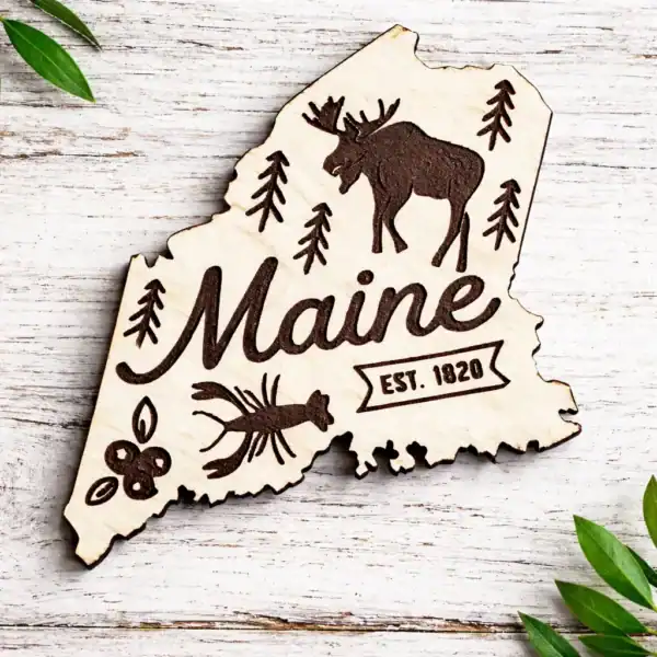 White Maine state–shaped wooden magnet with laser-engraved moose, pine trees, lobster, blueberries, and “Maine Est. 1820” design