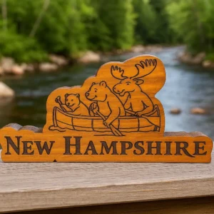 Engraved wooden New Hampshire sign featuring a moose, bear, and beaver paddling a canoe, displayed outdoors with a river in the background.