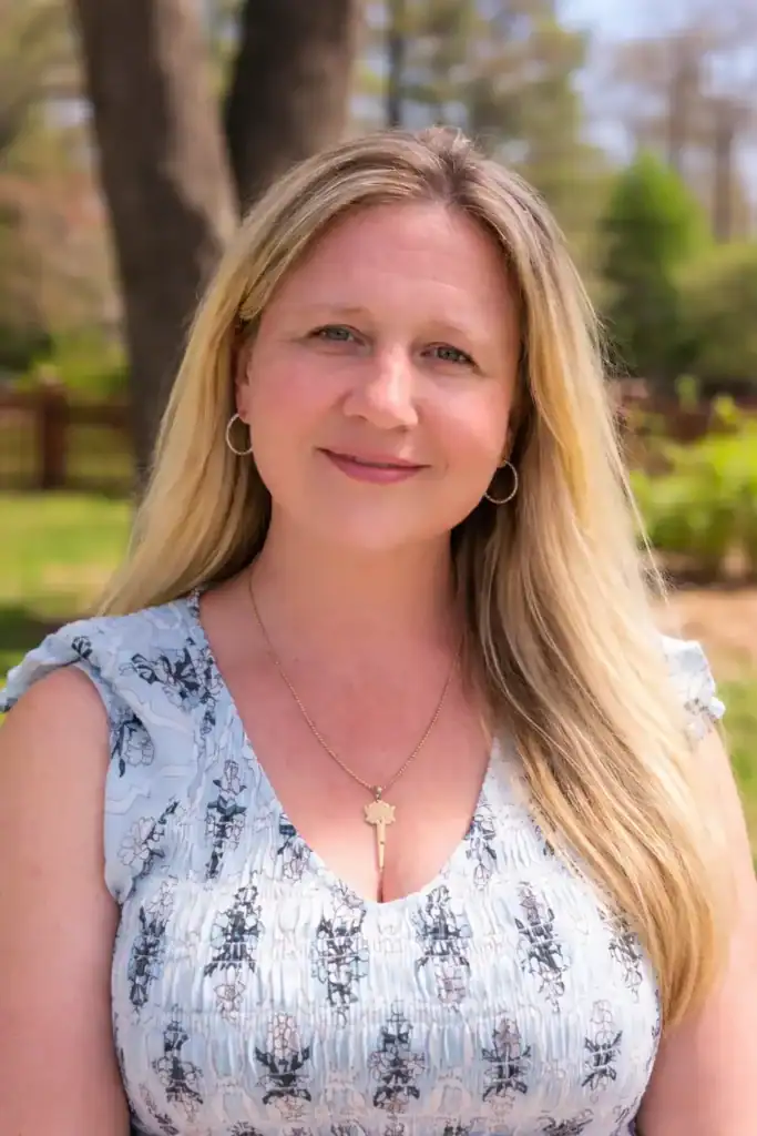 Kim Ross, co-founder of FMB Laser Designs, wearing a floral blouse and key pendant necklace—shot outdoors in natural light. A confident, approachable woman with long blonde hair, soft blue eyes, and a subtle smile, representing the leadership behind this woman-led New England studio.