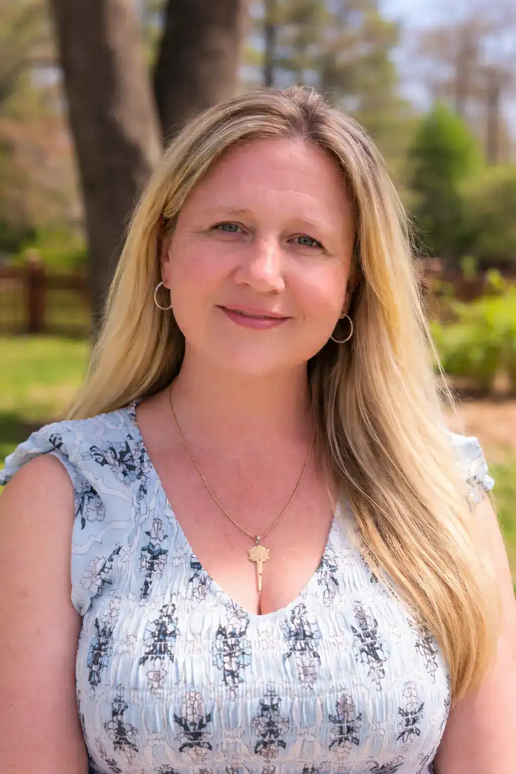 Kim Ross, co-founder of FMB Laser Designs, wearing a floral blouse and key pendant necklace—shot outdoors in natural light. A confident, approachable woman with long blonde hair, soft blue eyes, and a subtle smile, representing the leadership behind this woman-led New England studio.